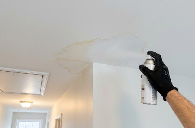 Gloved hand spraying stain-blocking primer from an aerosol can onto a water stain on a hallway ceiling
