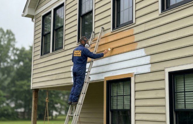 Painter on a ladder brushing primer onto replaced siding boards on a tan two-story house with black windows