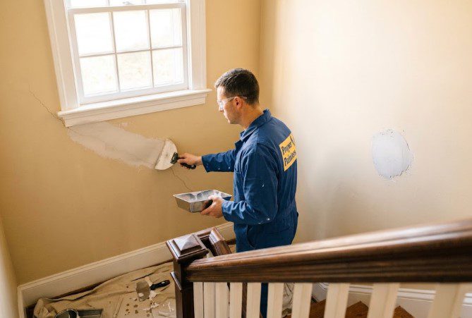 Painter filling a long plaster crack below a stairwell window with a putty knife and mud pan, with another patched spot nearby