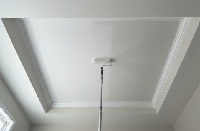 Painter rolling fresh white paint across a tray ceiling with an extension pole, with the new coat visibly brighter than the old